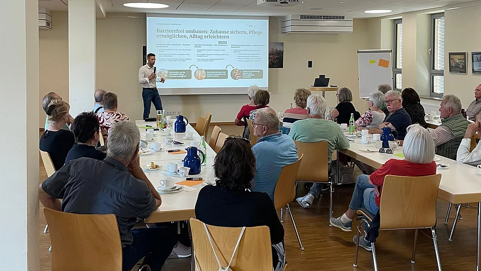 Ein Mann steht während seines Vortrags vor einer Präsentationsfläche, während zahlreiche Besucherinnen und Besucher an Tischen im Mehrgenerationenhaus sitzen und seinem Vortrag folgen.