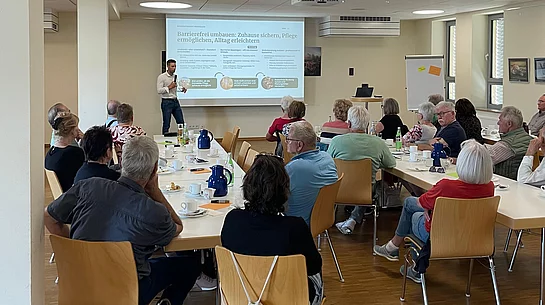 Ein Mann steht während seines Vortrags vor einer Präsentationsfläche, während zahlreiche Besucherinnen und Besucher an Tischen im Mehrgenerationenhaus sitzen und seinem Vortrag folgen.