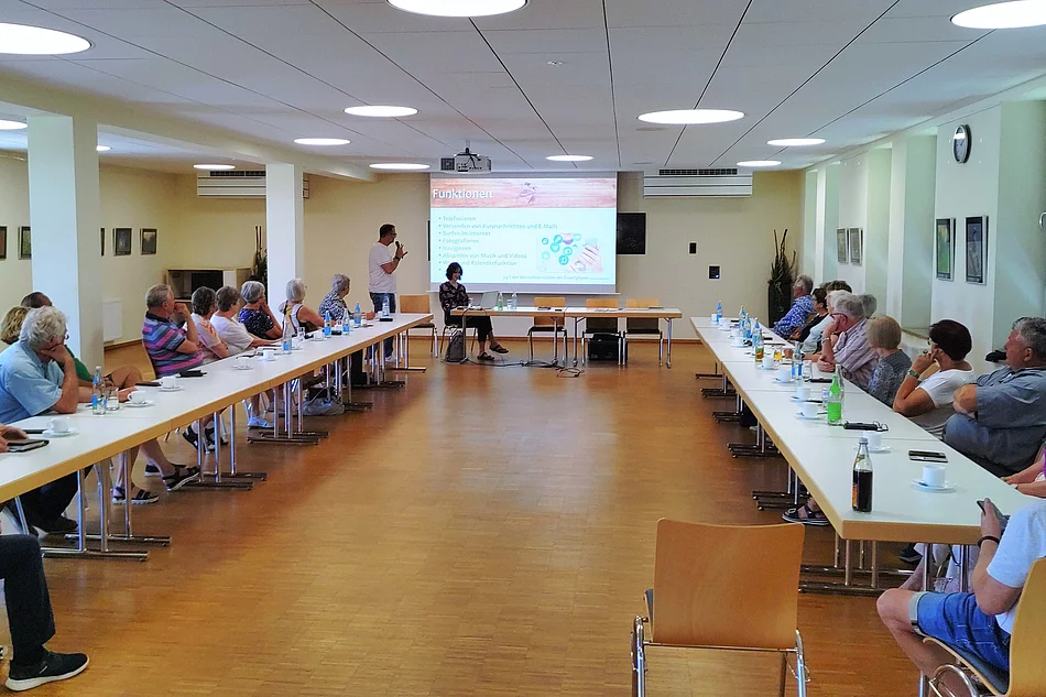 Mehrere ältere Menschen sitzen an Tischen in U-Form in einem hellen Raum und hören einem Vortrag zu, der mit einer Präsentation auf einer Leinwand unterstützt wird.