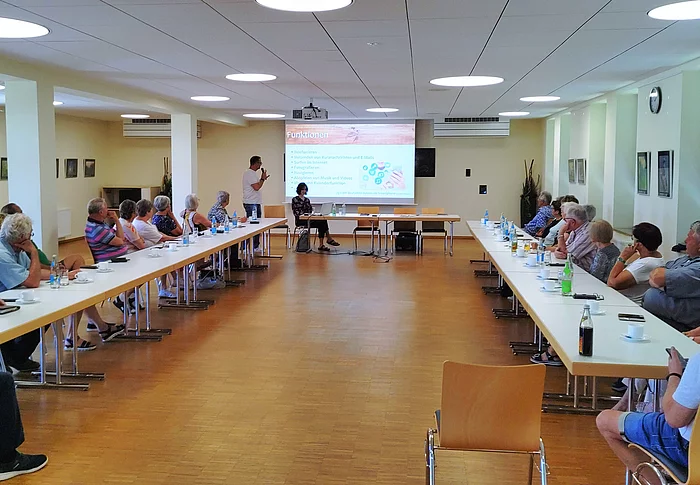 Mehrere ältere Menschen sitzen an Tischen in U-Form in einem hellen Raum und hören einem Vortrag zu, der mit einer Präsentation auf einer Leinwand unterstützt wird.