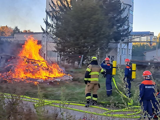 Mehrere Mitglieder der Jugendfeuerwehr und ein erwachsener Feuerwehrmann stehen mit Schläuchen auf einem Übungsgelände. Links brennt ein großer Haufen aus Holz und anderem Material mit hohen orangefarbenen Flammen, darüber steigt dunkler Rauch auf. Die Jugendlichen tragen dunkelblaue Schutzkleidung und rote Helme, der Feuerwehrmann vollständige Einsatzkleidung mit Helm. Das Foto entstand bei einer Feuerwehrübung im Rahmen des Sommerferienprogramms 2026.
