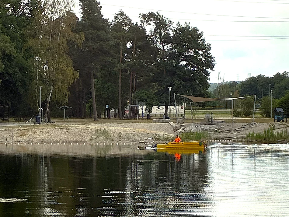 Ein gelbes Mähboot ist auf einem ruhigen Badesee im Einsatz und fährt nahe dem Ufer entlang. An Bord befindet sich eine Person in auffälliger orangefarbener Schutzkleidung. Im Hintergrund sind der Sandstrand der Badestelle, ein Beachvolleyballfeld, schattenspendende Bäume und weitere Freizeiteinrichtungen zu sehen. Das Boot entfernt Wasserpflanzen, um den Badebereich frei zu halten.