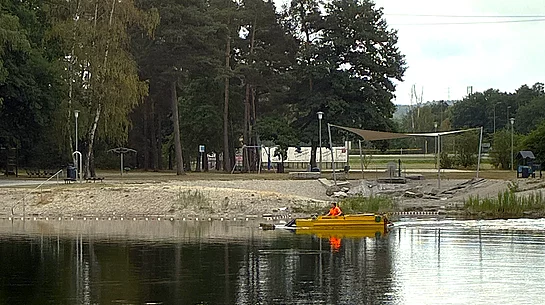 Ein gelbes Mähboot ist auf einem ruhigen Badesee im Einsatz und fährt nahe dem Ufer entlang. An Bord befindet sich eine Person in auffälliger orangefarbener Schutzkleidung. Im Hintergrund sind der Sandstrand der Badestelle, ein Beachvolleyballfeld, schattenspendende Bäume und weitere Freizeiteinrichtungen zu sehen. Das Boot entfernt Wasserpflanzen, um den Badebereich frei zu halten.