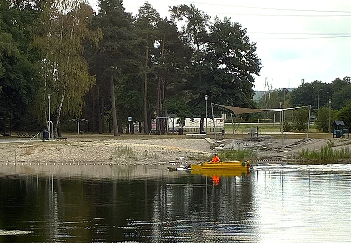Ein gelbes Mähboot ist auf einem ruhigen Badesee im Einsatz und fährt nahe dem Ufer entlang. An Bord befindet sich eine Person in auffälliger orangefarbener Schutzkleidung. Im Hintergrund sind der Sandstrand der Badestelle, ein Beachvolleyballfeld, schattenspendende Bäume und weitere Freizeiteinrichtungen zu sehen. Das Boot entfernt Wasserpflanzen, um den Badebereich frei zu halten.