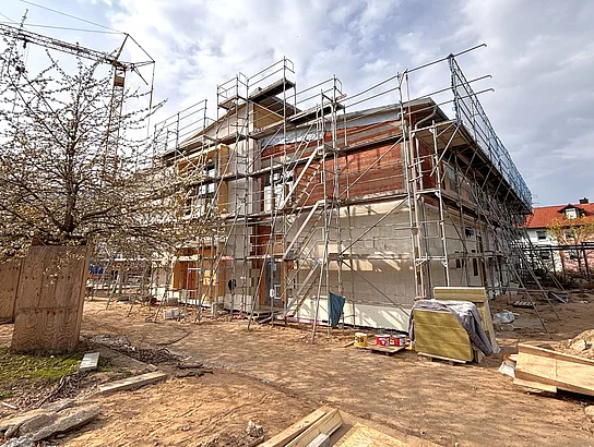 Ein Gebäude im Bau ist von Gerüsten umgeben, während Baumaterialien und Werkzeuge auf dem Gelände verteilt liegen.