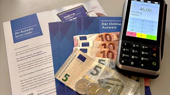 Auf dem Bild sieht man Informationsflyer zum Personalausweis, mehrere Euro-Geldscheine und Münzen sowie ein Kartenlesegerät, auf dessen Bildschirm ein zu zahlender Betrag angezeigt wird.