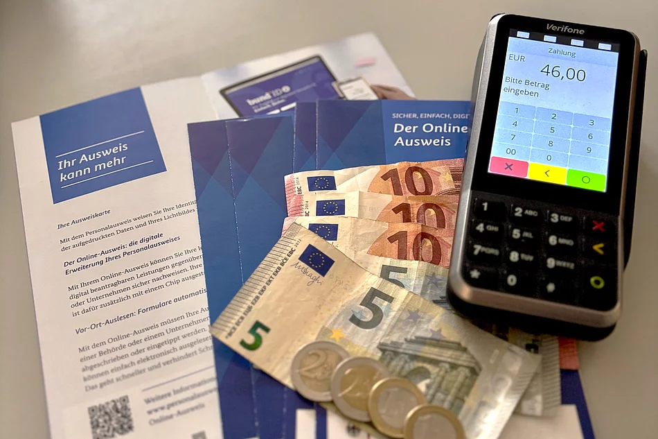 Auf dem Bild sieht man Informationsflyer zum Personalausweis, mehrere Euro-Geldscheine und Münzen sowie ein Kartenlesegerät, auf dessen Bildschirm ein zu zahlender Betrag angezeigt wird.