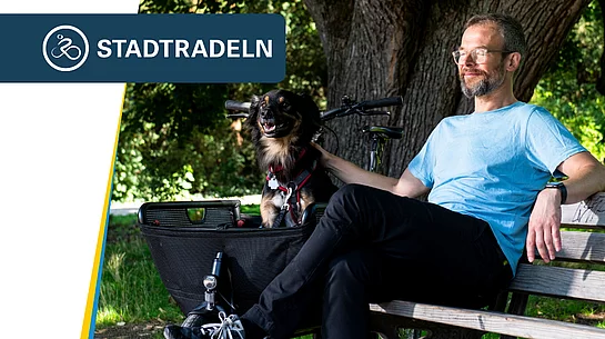 Ein Mann sitzt entspannt auf einer Parkbank im Schatten großer Bäume. Neben ihm befindet sich ein Fahrrad mit einem Korb, in dem ein kleiner schwarz-brauner Hund sitzt. Der Mann trägt ein hellblaues T-Shirt und blickt zufrieden zur Seite, während er den Hund streichelt. Oben links ist das Logo der Aktion „STADTRADELN“ eingeblendet.