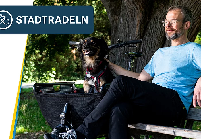 Ein Mann sitzt entspannt auf einer Parkbank im Schatten großer Bäume. Neben ihm befindet sich ein Fahrrad mit einem Korb, in dem ein kleiner schwarz-brauner Hund sitzt. Der Mann trägt ein hellblaues T-Shirt und blickt zufrieden zur Seite, während er den Hund streichelt. Oben links ist das Logo der Aktion „STADTRADELN“ eingeblendet.