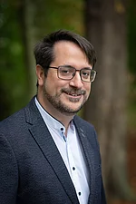Ein Mann mit Brille und Bart trägt ein Sakko und schaut freundlich zur Seite.