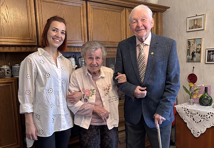 Eine jüngere Frau steht lächelnd neben einem älteren Ehepaar in einem Wohnzimmer, während die Frau den Arm der Seniorin hält und der Mann einen Gehstock in der Hand hat.