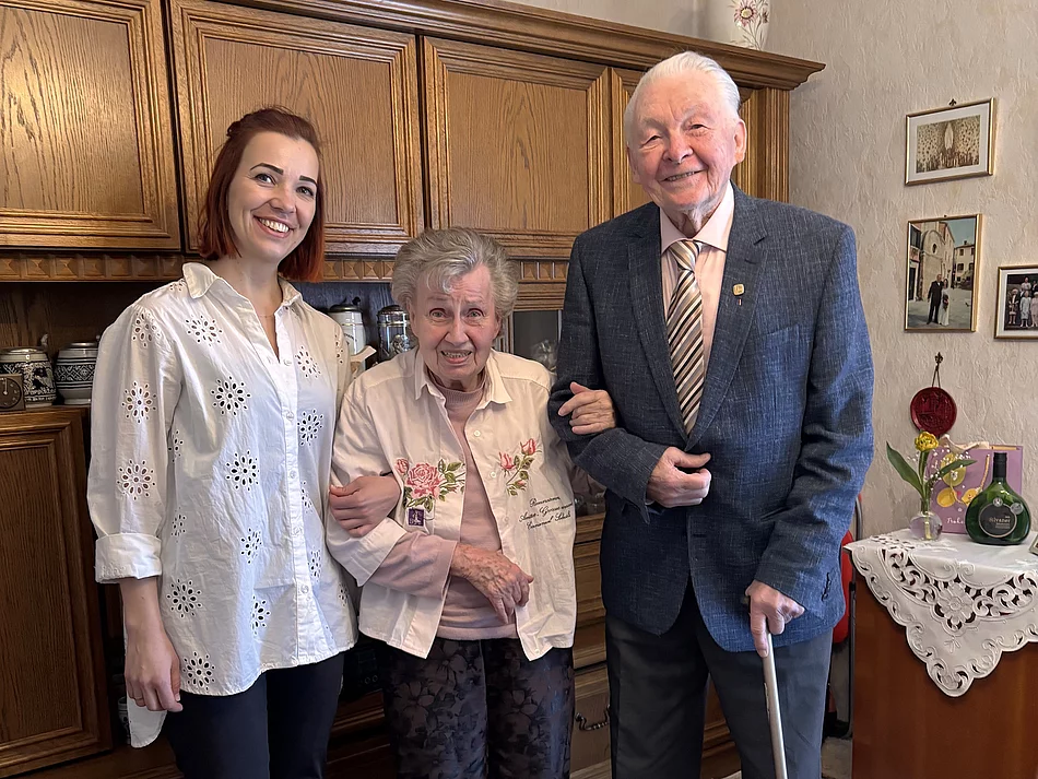 Eine jüngere Frau steht lächelnd neben einem älteren Ehepaar in einem Wohnzimmer, während die Frau den Arm der Seniorin hält und der Mann einen Gehstock in der Hand hat.