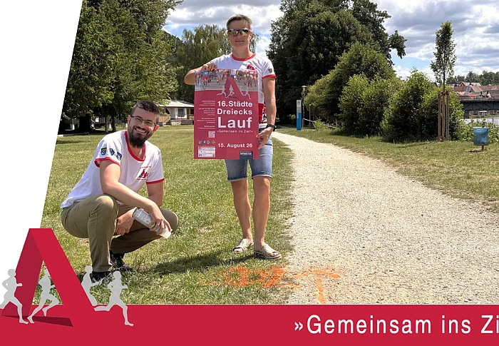Eine Werbegrafik zeigt zwei Personen in Laufshirts des Städtedreieckslaufs an einem geschotterten Uferweg. Eine Person kniet neben einer orangefarbenen Streckenmarkierung auf dem Boden, die andere hält das Veranstaltungsplakat des 16. Städtedreieckslaufs in die Kamera. Im Hintergrund sind Bäume, die Naab sowie eine Brücke zu sehen. Am unteren Bildrand steht der Slogan „Gemeinsam ins Ziel!“. Die Grafik weist darauf hin, dass die Laufstrecken bereits markiert sind und zum Training genutzt werden können.