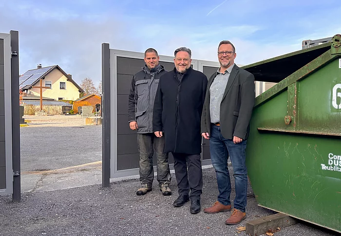 Drei Männer stehen lächelnd neben einem großen grünen Container auf einem Hof mit offenem Tor; im Hintergrund sind ein Wohnhaus mit Solardach und ein Friedhof zu sehen.
