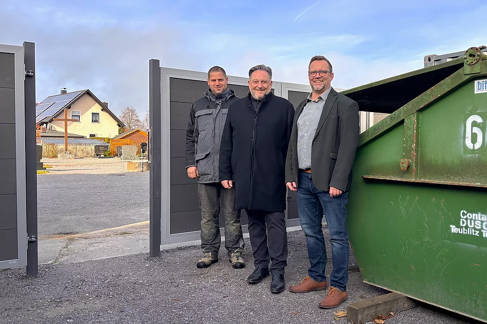 Drei Männer stehen lächelnd neben einem großen grünen Container auf einem Hof mit offenem Tor; im Hintergrund sind ein Wohnhaus mit Solardach und ein Friedhof zu sehen.