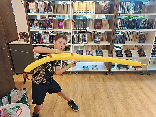 Ein Junge steht vor Bücherregalen in der Stadtbücherei und hält einen großen selbst gebauten gelben Bogen mit gespannter Schnur in den Händen.