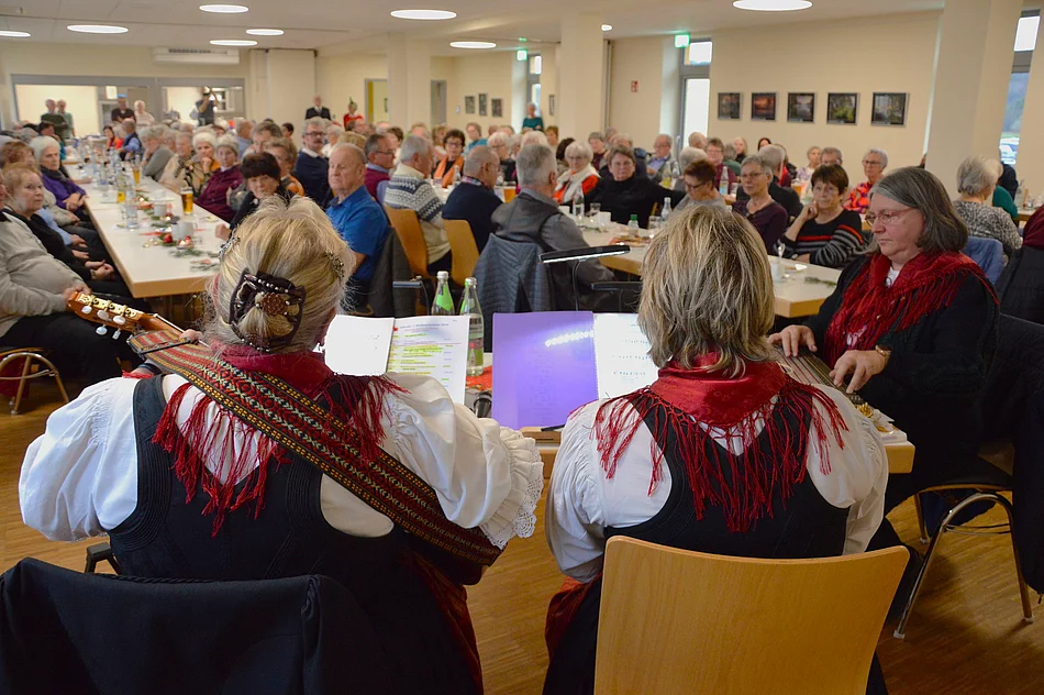 Musikerinnen in traditioneller Kleidung spielen vor zahlreichen Seniorinnen und Senioren, die an festlich gedeckten Tischen einer Adventsfeier lauschen.