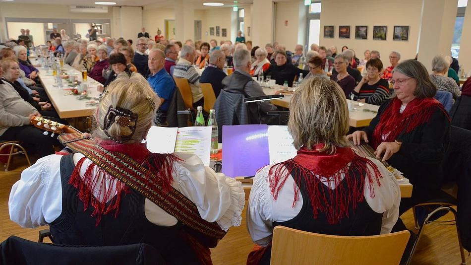 Musikerinnen in traditioneller Kleidung spielen vor zahlreichen Seniorinnen und Senioren, die an festlich gedeckten Tischen einer Adventsfeier lauschen.