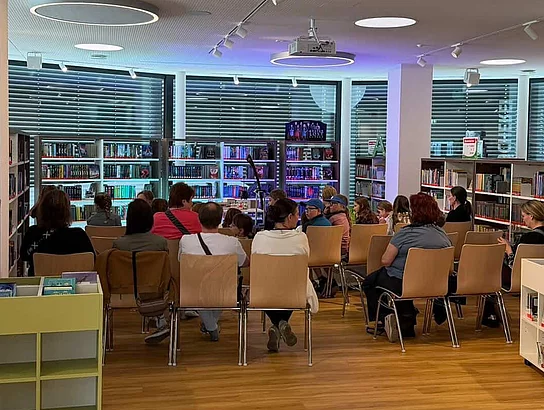 Mehrere Kinder und Erwachsene sitzen in einer modern eingerichteten Bücherei auf Stühlen und verfolgen aufmerksam eine Veranstaltung. Im Hintergrund sind Bücherregale mit stimmungsvoller farbiger Beleuchtung zu sehen. Die Atmosphäre wirkt ruhig und gespannt.