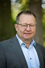 Ein Mann mittleren Alters mit Brille und grauem Anzug schaut ruhig und direkt in die Kamera.