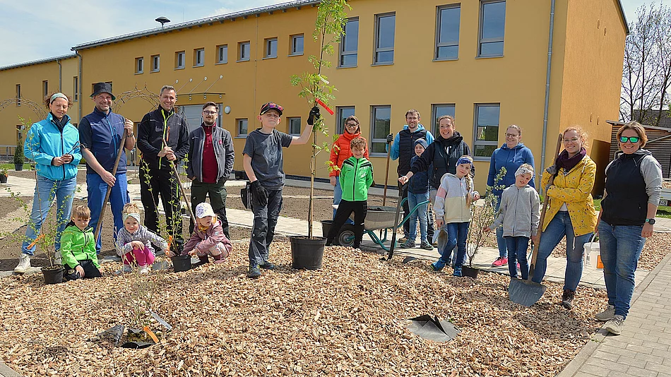 Eine Gruppe fröhlich grinsender Menschen von Jung bis Alt bei der Anpflanzaktion der Wildkräuterwiese. Im Hintergrund das orange-gelbe Gebäude des Mehrgenerationenhauses.