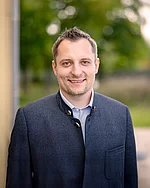 Ein Mann mit kurzem Haar und traditionell wirkender Jacke lächelt leicht.