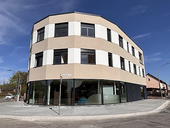 Außenansicht eines modernen, mehrstöckigen Gebäudes mit großen Glasflächen an einer Straßenecke.