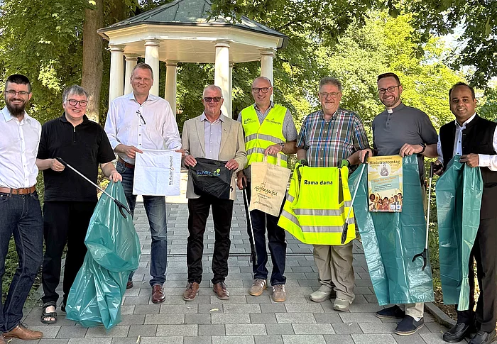 Eine Gruppe aus Vertreterinnen und Vertretern der Städte, Organisatoren sowie kirchlichen Gemeinden des Städtedreiecks steht vor einem Pavillon im Stadtpark. Mehrere Personen halten Müllsäcke, Greifzangen, Warnwesten oder Informationsmaterialien in den Händen und werben gemeinsam für die bevorstehende Rama-Dama-Aufräumaktion.
