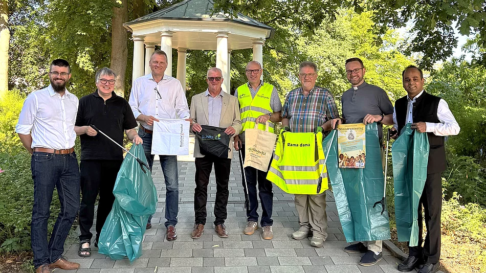 Eine Gruppe aus Vertreterinnen und Vertretern der Städte, Organisatoren sowie kirchlichen Gemeinden des Städtedreiecks steht vor einem Pavillon im Stadtpark. Mehrere Personen halten Müllsäcke, Greifzangen, Warnwesten oder Informationsmaterialien in den Händen und werben gemeinsam für die bevorstehende Rama-Dama-Aufräumaktion.