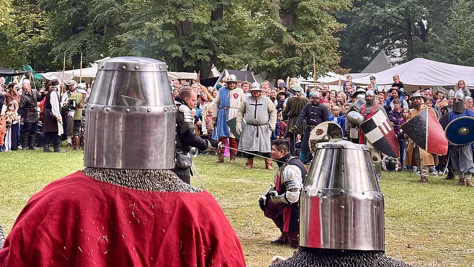 Im Vordergrund stehen zwei Ritter in Kettenhemd und Topfhelm mit dem Rücken zur Kamera und beobachten das Kampfgeschehen auf einer großen Wiese. In der Mitte des Bildes kniet ein weiterer Ritter in Rüstung, während sich weitere Kämpfer mit Schwertern und Schilden gegenüberstehen. Hinter dem Kampf versammeln sich zahlreiche Besucherinnen und Besucher, die das Spektakel verfolgen. Mittelalterliche Lagerzelte und hohe Bäume bilden die Kulisse des Stadtparks.
