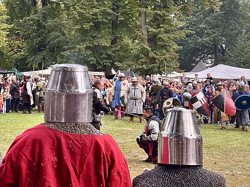 Im Vordergrund stehen zwei Ritter in Kettenhemd und Topfhelm mit dem Rücken zur Kamera und beobachten das Kampfgeschehen auf einer großen Wiese. In der Mitte des Bildes kniet ein weiterer Ritter in Rüstung, während sich weitere Kämpfer mit Schwertern und Schilden gegenüberstehen. Hinter dem Kampf versammeln sich zahlreiche Besucherinnen und Besucher, die das Spektakel verfolgen. Mittelalterliche Lagerzelte und hohe Bäume bilden die Kulisse des Stadtparks.