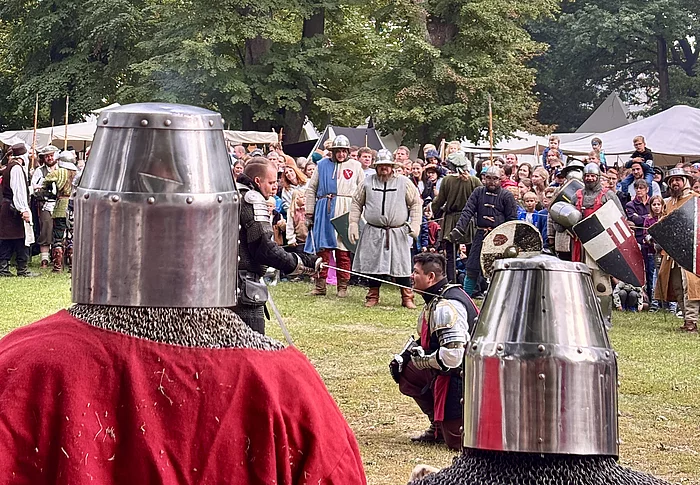 Im Vordergrund stehen zwei Ritter in Kettenhemd und Topfhelm mit dem Rücken zur Kamera und beobachten das Kampfgeschehen auf einer großen Wiese. In der Mitte des Bildes kniet ein weiterer Ritter in Rüstung, während sich weitere Kämpfer mit Schwertern und Schilden gegenüberstehen. Hinter dem Kampf versammeln sich zahlreiche Besucherinnen und Besucher, die das Spektakel verfolgen. Mittelalterliche Lagerzelte und hohe Bäume bilden die Kulisse des Stadtparks.