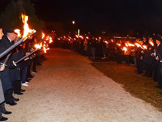 Zwei Reihen von Menschen stehen sich nachts gegenüber und bilden mit brennenden Fackeln einen leuchtenden Ehrenbogen entlang eines Kieswegs.