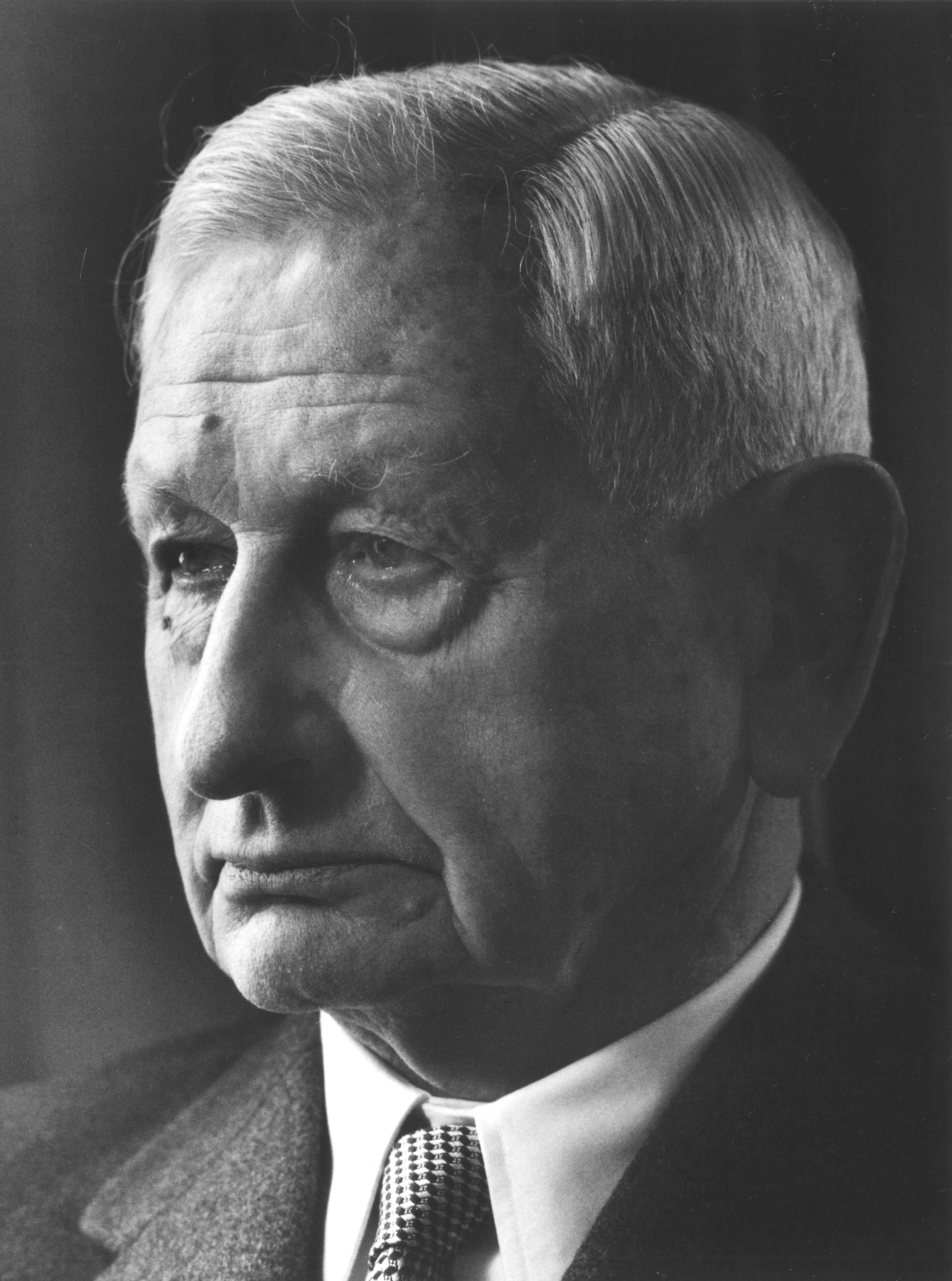Das Schwarz-Weiß-Foto zeigt einen älteren Mann im Anzug. Er hat kurze, glatt nach hinten gekämmte, graue Haare und ein markantes Gesicht mit tieferen Falten. Sein Blick ist ernst und nachdenklich, er schaut leicht zur Seite. Das Gesicht wirkt streng, die Lippen sind geschlossen. Er trägt ein weißes Hemd mit Krawatte und ein dunkles Jackett. Die Beleuchtung fällt von vorne, sodass die Gesichtszüge deutlich hervorgehoben sind.