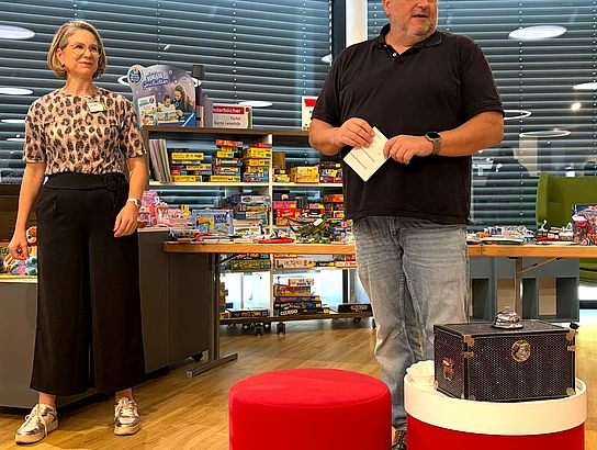 Büchereileiterin Corinna Höfler und Bürgermeister Thomas Beer stehen vor einem Tisch mit zahlreichen Preisen in der Stadtbücherei; Thomas Beer hält einen Zettel in den Händen.