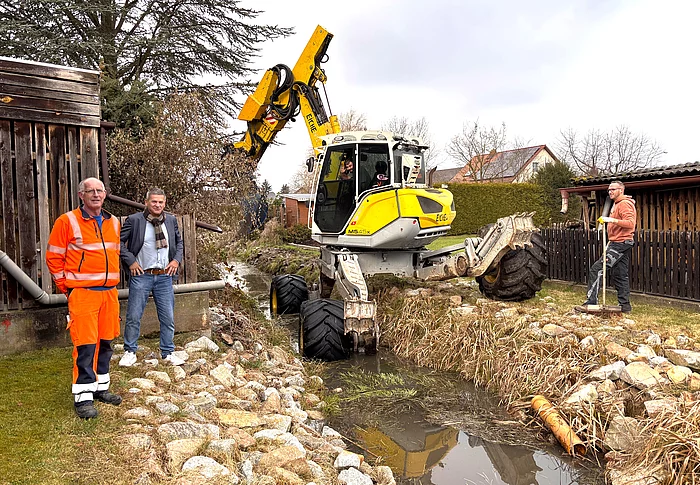 Ein Schreitbagger arbeitet am Ufer eines kleinen Grabens, während drei Männer – einer in Arbeitskleidung, zwei in Zivil – daneben stehen und die Maßnahmen beobachten.