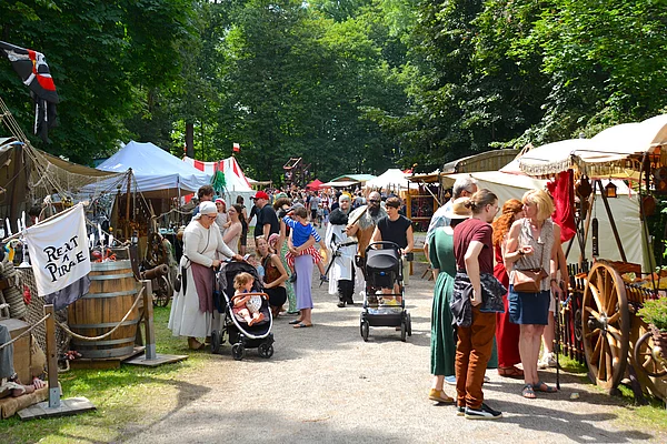 Eine belebte Marktgasse des Mittelalterfestes im Stadtpark Teublitz. Links und rechts säumen mittelalterliche Händlerstände und Zelte den Weg. Zahlreiche Besucherinnen und Besucher schlendern durch den Markt, viele Familien mit Kindern und Kinderwagen. Zwischen den Ständen sind historische Gewandungen, Handwerkswaren und Dekorationen zu sehen. Hohe Bäume spenden Schatten und verleihen dem Gelände eine besondere Atmosphäre.