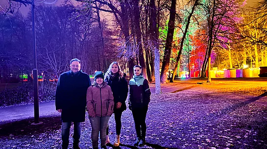 Vier Personen stehen abends im farbig bunt beleuchteten Stadtpark, während Lampen und angestrahlte Bäume eine festliche Atmosphäre erzeugen.