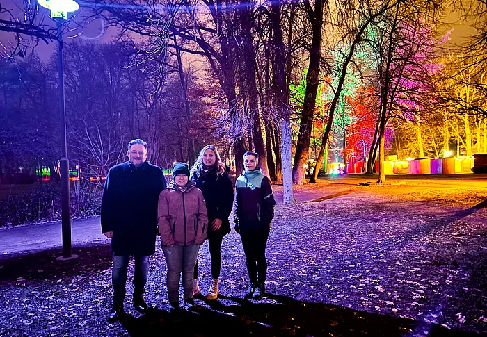 Vier Personen stehen abends im farbig bunt beleuchteten Stadtpark, während Lampen und angestrahlte Bäume eine festliche Atmosphäre erzeugen.