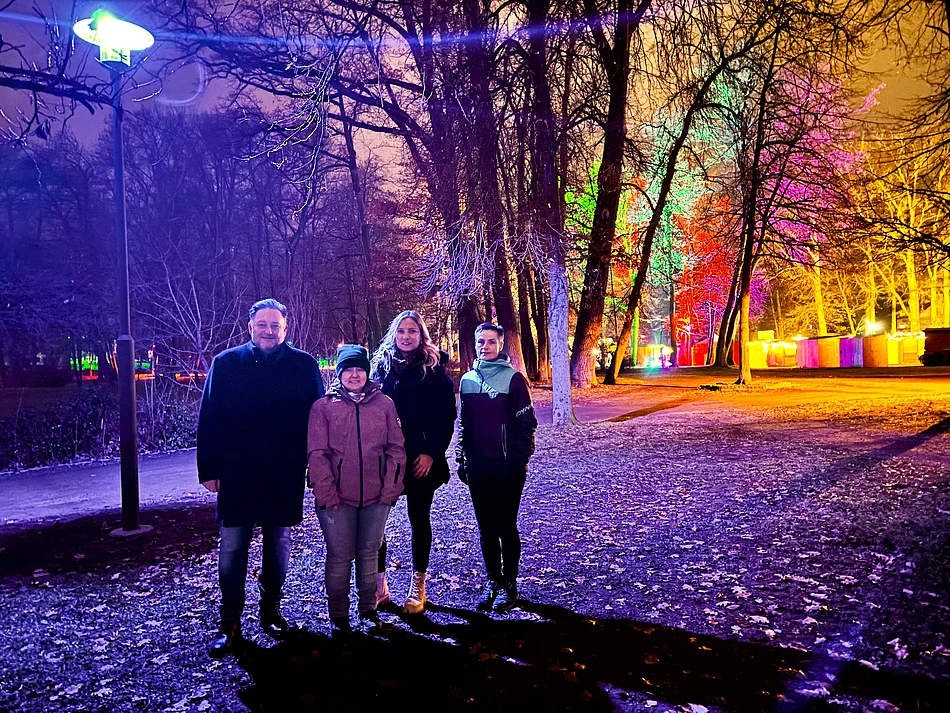 Vier Personen stehen abends im farbig bunt beleuchteten Stadtpark, während Lampen und angestrahlte Bäume eine festliche Atmosphäre erzeugen.