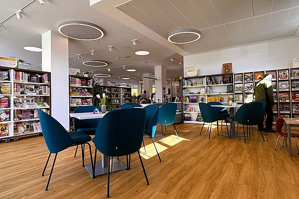 Offener Aufenthaltsbereich der Stadtbücherei mit blauen Sesseln und Tischen. Umgeben von Bücherregalen bietet der Raum Platz zum Lesen, Lernen oder Austauschen. Eine Kaffee-Station und verschiedene Sitzgelegenheiten schaffen Café-Atmosphäre.