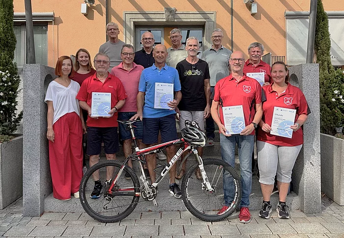 Das Foto zeigt eine Gruppe von 15 Menschen, die gemeinsam vor einem Gebäude mit orangener Fassade stehen. Im Vordergrund, in der Mitte, steht ein Mountainbike. Davor und daneben stehen fünf Personen, die Urkunden in den Händen halten. Vier von ihnen tragen rote Shirts mit einem Vereinswappen, eine Person trägt ein hellblaues Polohemd. Rechts vorne steht eine Frau in rotem Shirt und weißen Hosen, die ebenfalls eine Urkunde hält. Die Gruppe wirkt fröhlich, viele lächeln. Im Hintergrund stehen weitere Personen ohne Urkunden. Das Gebäude hinter ihnen hat einen überdachten Eingang mit Glasdach, flankiert von großen Pflanzkübeln mit Bäumen und weißen Blumen.