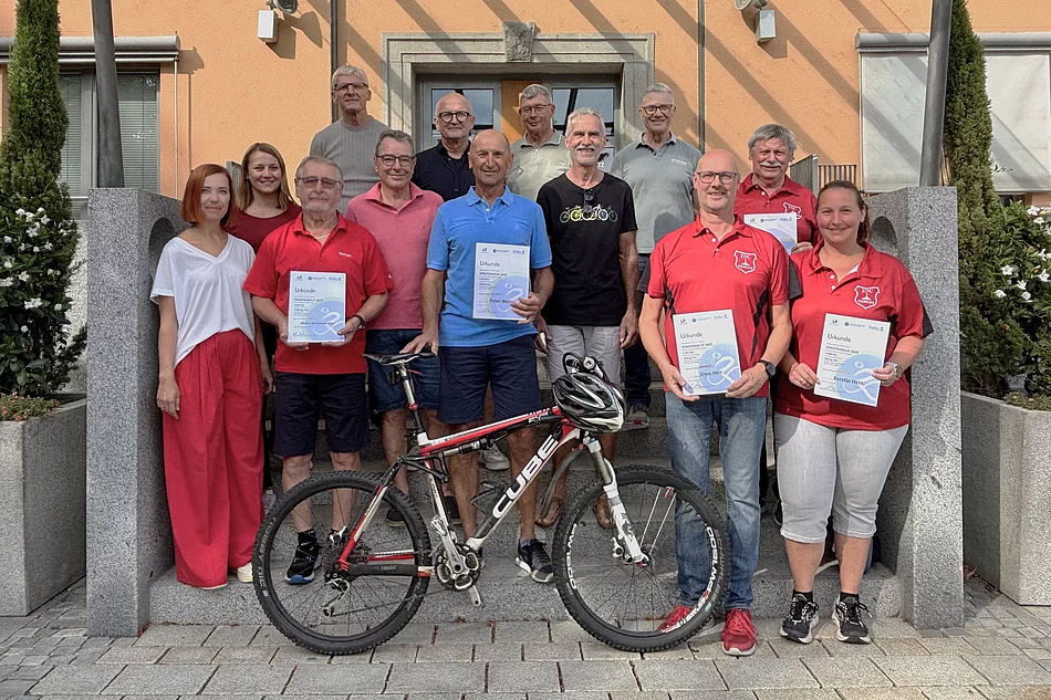 Das Foto zeigt eine Gruppe von 15 Menschen, die gemeinsam vor einem Gebäude mit orangener Fassade stehen. Im Vordergrund, in der Mitte, steht ein Mountainbike. Davor und daneben stehen fünf Personen, die Urkunden in den Händen halten. Vier von ihnen tragen rote Shirts mit einem Vereinswappen, eine Person trägt ein hellblaues Polohemd. Rechts vorne steht eine Frau in rotem Shirt und weißen Hosen, die ebenfalls eine Urkunde hält. Die Gruppe wirkt fröhlich, viele lächeln. Im Hintergrund stehen weitere Personen ohne Urkunden. Das Gebäude hinter ihnen hat einen überdachten Eingang mit Glasdach, flankiert von großen Pflanzkübeln mit Bäumen und weißen Blumen.