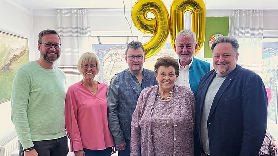 Sechs Personen stehen lächelnd nebeneinander in einem hellen Raum, vor ihnen eine ältere Frau im Mittelpunkt, während im Hintergrund zwei große goldene Luftballons mit der Zahl „90“ an der Decke hängen.