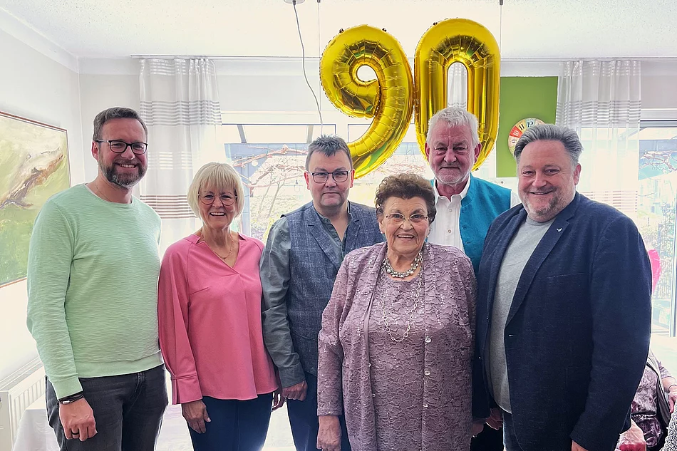 Sechs Personen stehen lächelnd nebeneinander in einem hellen Raum, vor ihnen eine ältere Frau im Mittelpunkt, während im Hintergrund zwei große goldene Luftballons mit der Zahl „90“ an der Decke hängen.