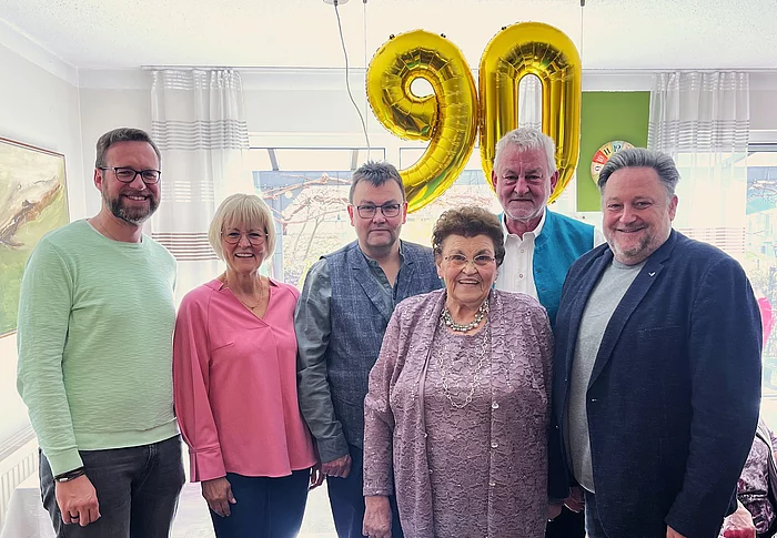 Sechs Personen stehen lächelnd nebeneinander in einem hellen Raum, vor ihnen eine ältere Frau im Mittelpunkt, während im Hintergrund zwei große goldene Luftballons mit der Zahl „90“ an der Decke hängen.