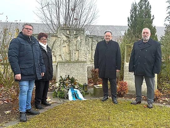 Vier Menschen stehen bei Tageslicht vor einem steinernen Kriegerdenkmal mit einem niedergelegten Kranz.