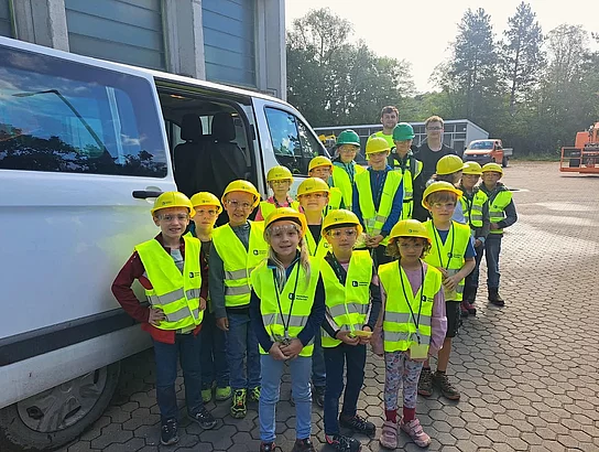 Eine Gruppe von Kindern steht gemeinsam mit zwei erwachsenen Begleitpersonen auf einem Betriebsgelände vor einem weißen Kleinbus. Die Kinder tragen leuchtend gelbe Warnwesten sowie überwiegend gelbe, teilweise grüne Schutzhelme und Schutzbrillen. Im Hintergrund sind Betriebsgebäude, Fahrzeuge und Bäume zu sehen.