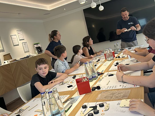Kinder und Erwachsene sitzen an einem langen Tisch und bauen kleine Fahrzeuge zusammen. Ein Betreuer erklärt vorne einen Arbeitsschritt; auf dem Tisch liegen Bauteile, Werkzeuge und Arbeitsunterlagen.