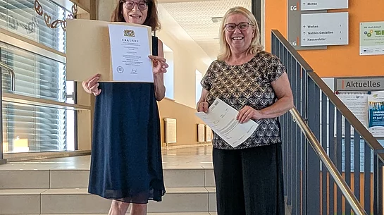 Zwei Frauen stehen lächelnd auf einer Treppe im Eingangsbereich der Telemann-Grundschule. Petra Koch hält ihre Pensionsurkunde in den Händen, daneben steht Schulleiterin Sabine Kunz mit weiteren Unterlagen.